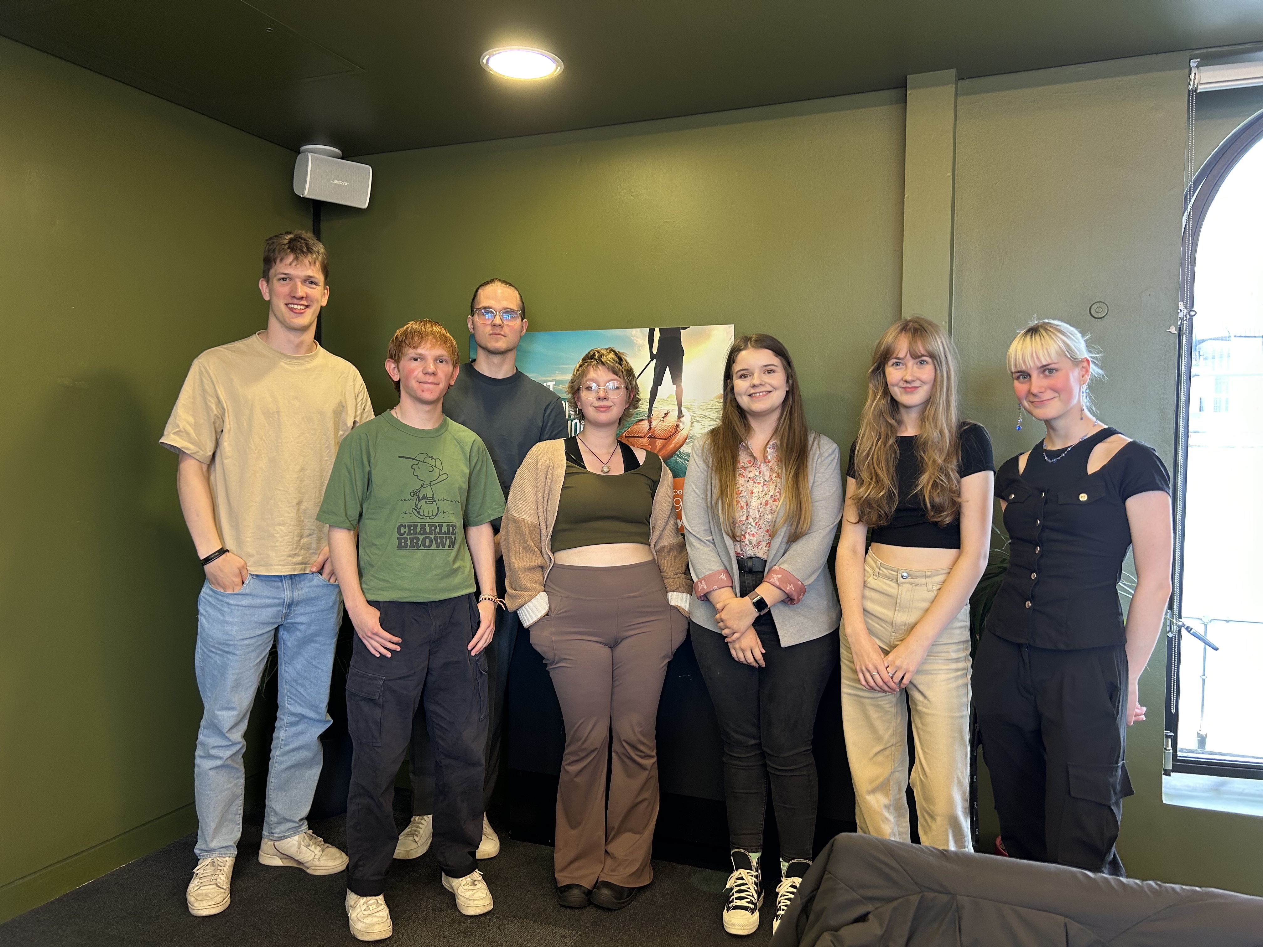 Seven young adults standing indoors against a green wall, smiling at the camera.