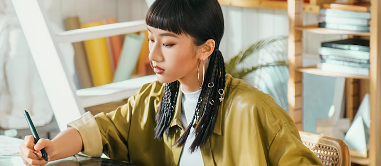 Young woman with braided hair and hoop earrings focused on writing at a desk in a sunlit room with books and plants.