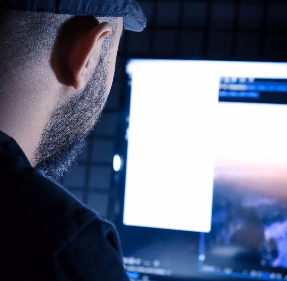 Rear view of a man with a beard and cap working on a computer with bright screens.