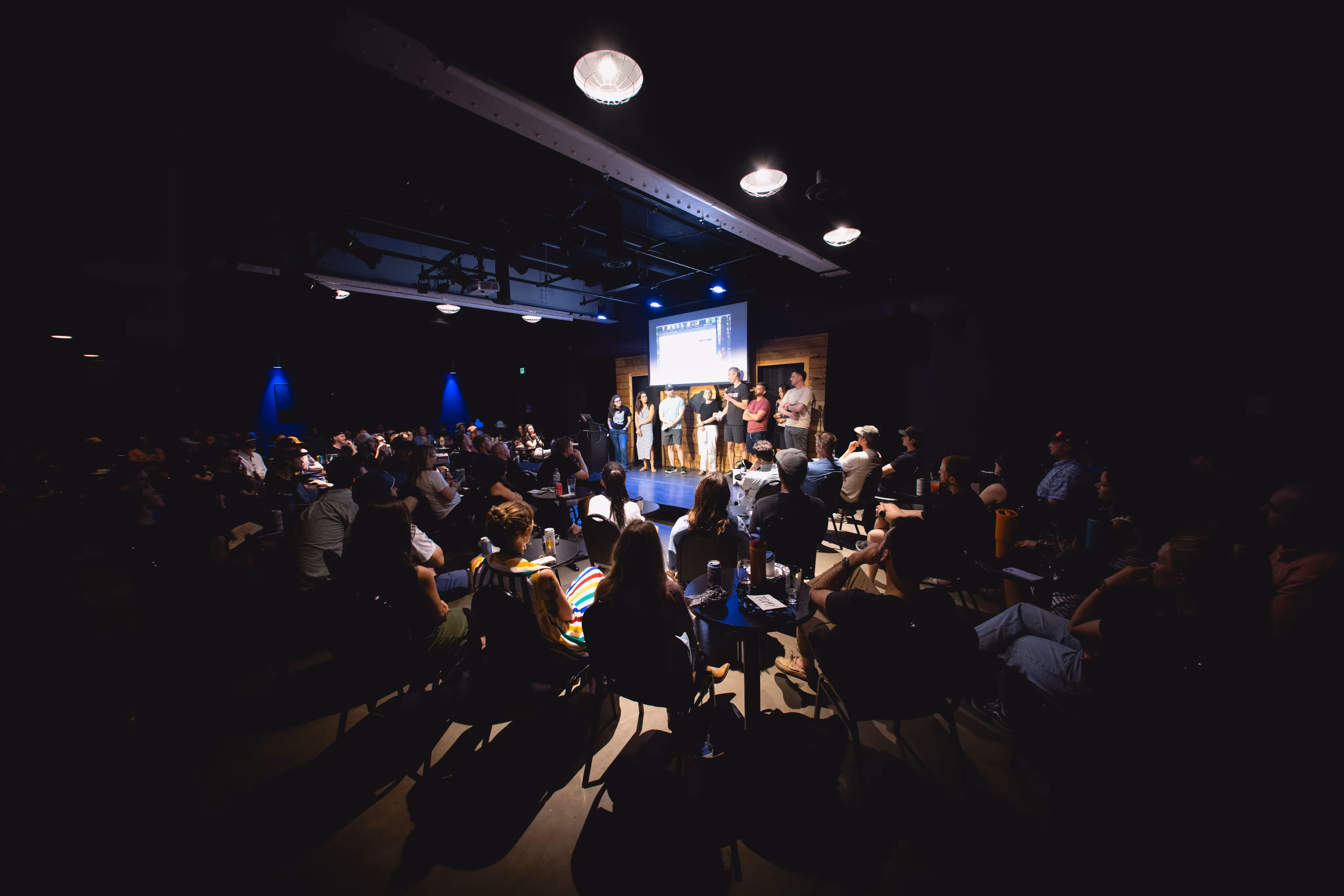 Audience seated in a dark room watching a group of people standing on a lit stage with a presentation screen behind them.