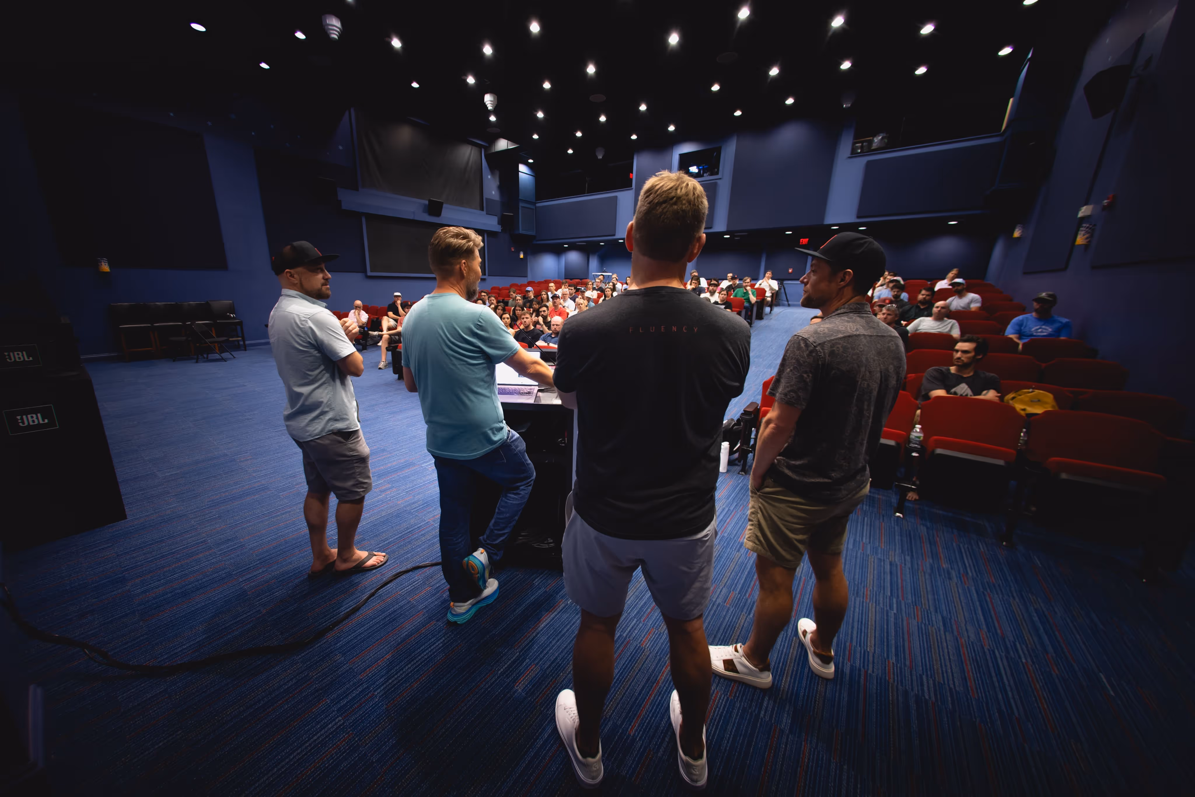 Four men standing in front of a podium addressing an audience seated in a theater-style room.