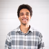 Smiling young man with curly hair wearing a gray plaid shirt against a white background.