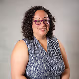 Smiling woman with curly hair and glasses wearing a sleeveless patterned top against a light gray background.
