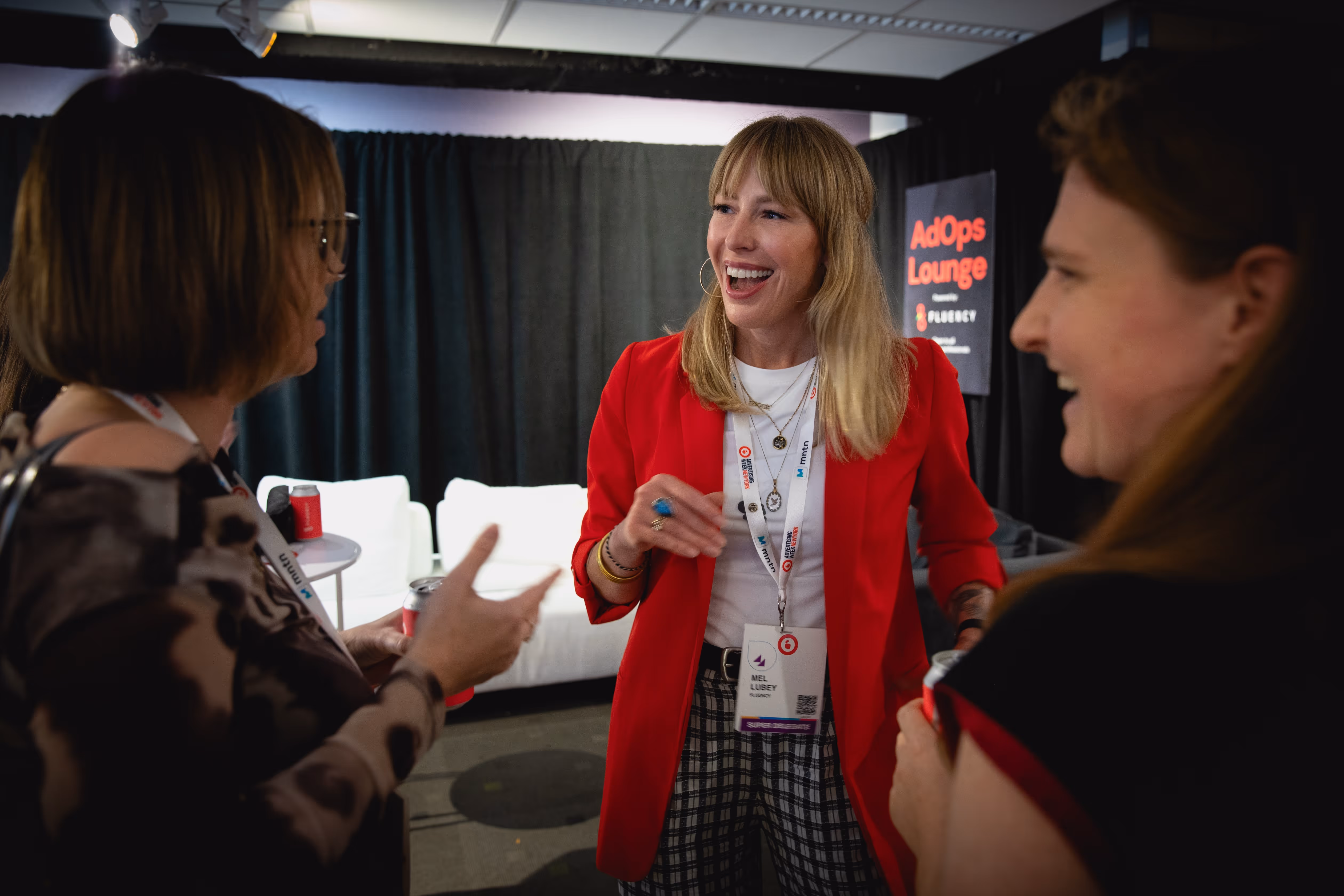 Three women engaged in lively conversation at an indoor event, with one woman in a red blazer smiling broadly.