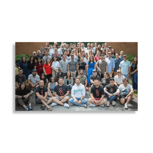 Large group of diverse adults posing outdoors in front of a brick wall, smiling at the camera.