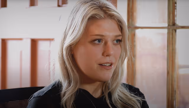 Blonde woman speaking indoors with wooden window and door in the background.