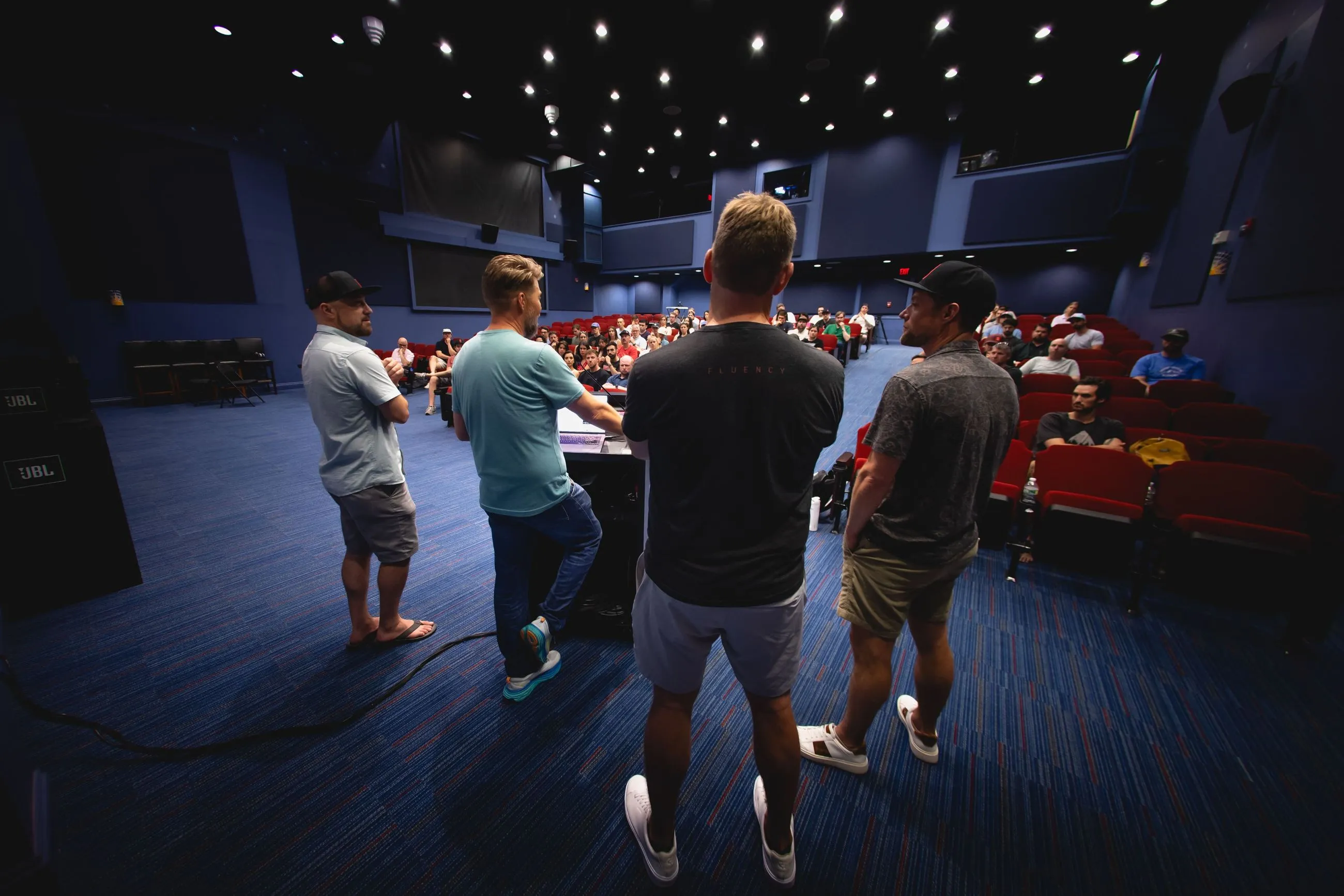 Four men standing and speaking to an audience seated in a dark auditorium with red chairs.