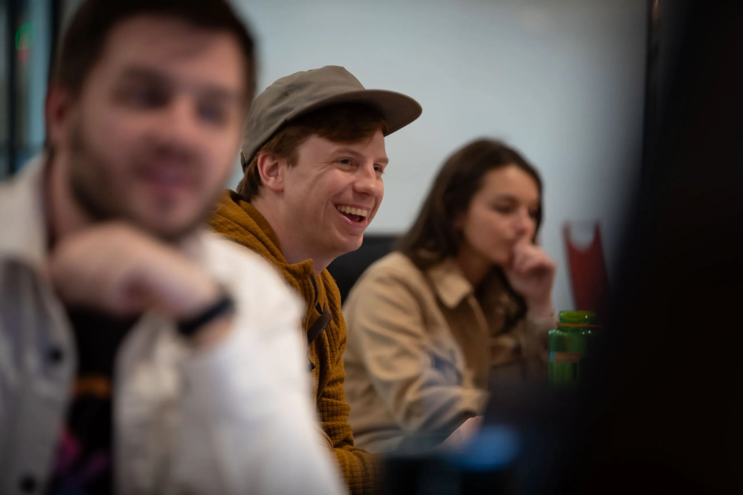 Three young adults in a casual setting, with one smiling man wearing a cap in focus in the middle.