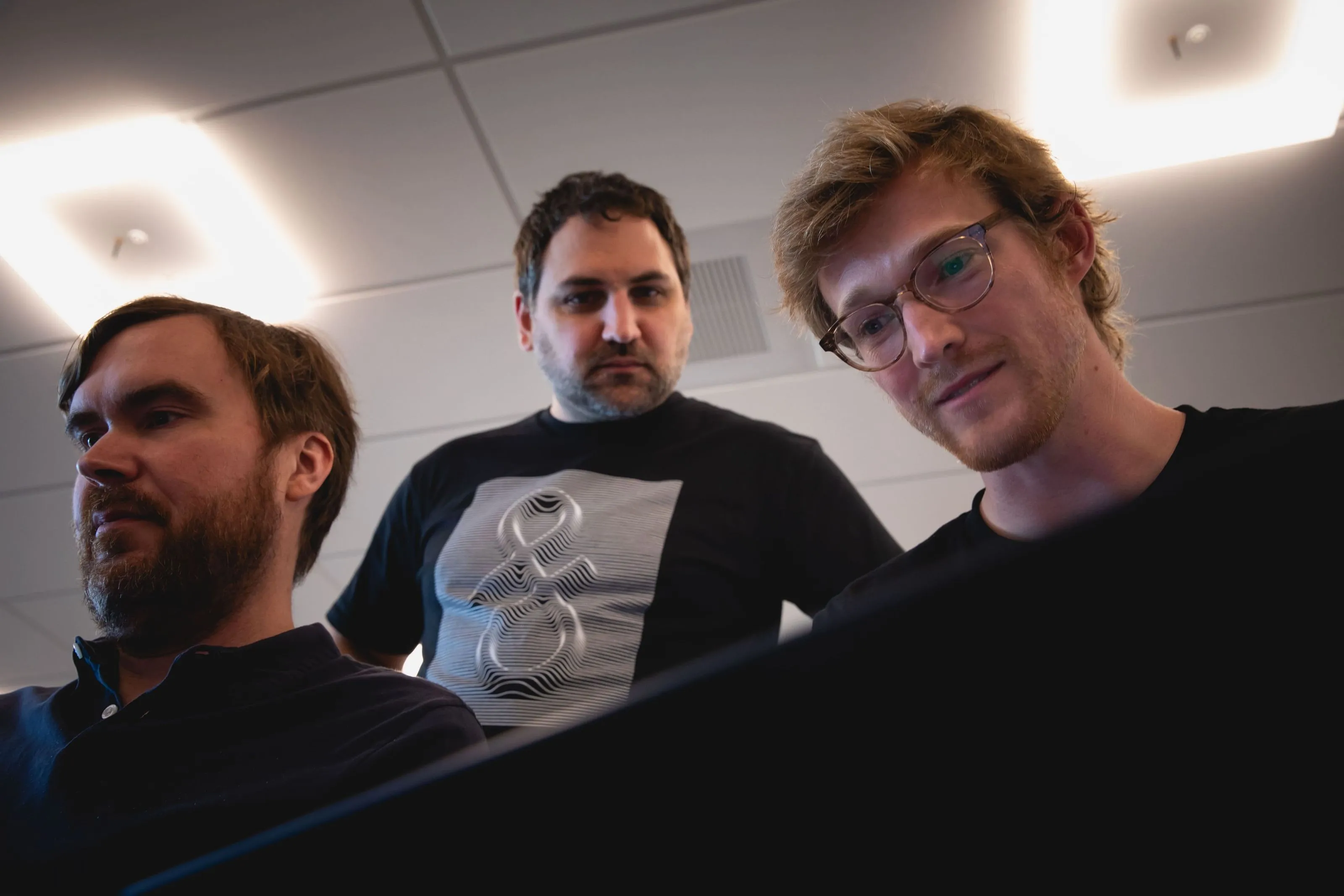 Three men attentively looking at a laptop screen indoors under office ceiling lights.