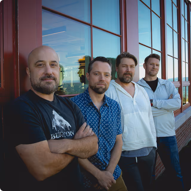 Four men standing side by side with arms crossed, leaning against a windowed wall outside.