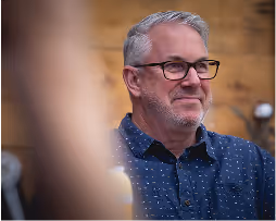 Middle-aged man with gray hair and glasses smiling while wearing a blue patterned shirt.