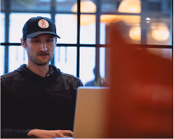 Man wearing a black cap with a white logo working on a laptop in front of a window.