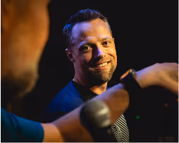 Smiling man with short hair and a striped shirt speaking into a microphone, lit against a dark background.