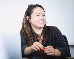 Smiling woman with long dark hair sitting and holding a pen, wearing a black blouse.