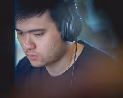 Young man wearing headphones, focused on something off-screen in a dimly lit setting.