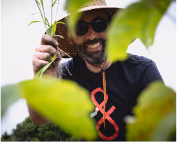 Smiling man wearing sunglasses and a hat holds a green plant among large leaves.