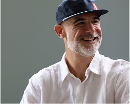 Smiling middle-aged man with white beard wearing a black baseball cap and white shirt against a gray background.