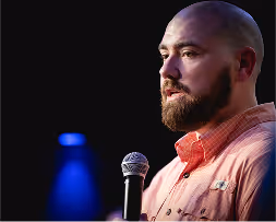 Bearded man in an orange shirt speaking into a microphone on a dark stage with blue spotlight in background.