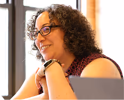 Curly-haired woman wearing glasses smiling and resting her chin on her hands near a laptop.