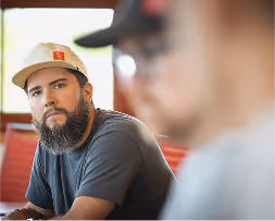 Man with beard and beige cap looking attentively while blurred person is in the foreground.
