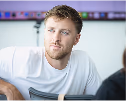 Young man with light brown hair and beard looking to the side wearing a white shirt indoors.