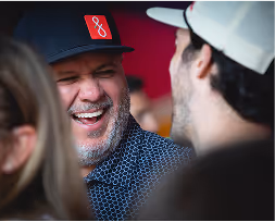 Man with gray beard wearing a black cap with red and white logo smiling and talking to another person in a social setting.