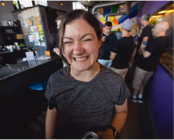 Smiling woman with dark hair in a gray t-shirt at a busy indoor venue with people in the background.