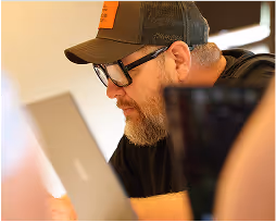 Bearded man wearing glasses and a cap focused on a laptop screen.
