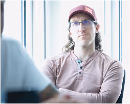 Man wearing glasses and a red cap sitting with arms crossed near a large window.