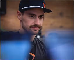 Man with a mustache wearing a black cap and shirt looking down in an indoor setting.
