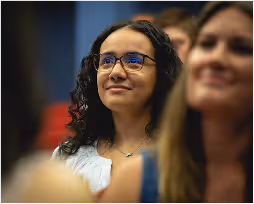 Woman with curly hair and glasses attending an event, smiling attentively.