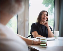 Smiling woman sitting at a table with a cup of coffee, engaging in conversation with a blurred person in the foreground.