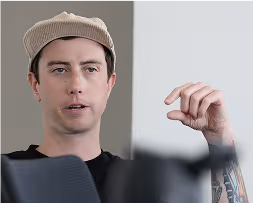 Man wearing a beige cap and black shirt gesturing with his hand while speaking.