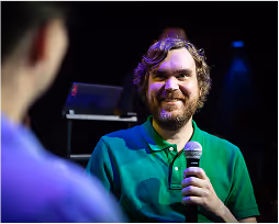 Bearded man in green shirt holding a microphone and smiling during a conversation on stage.