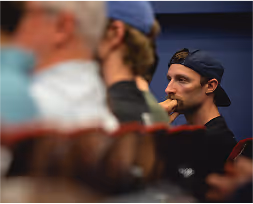 Man wearing a backward cap, biting his nails and looking focused in a dimly lit indoor setting.