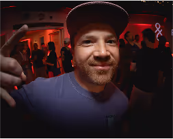 Smiling man in a cap pointing his finger at a dimly lit party with red and orange lighting.