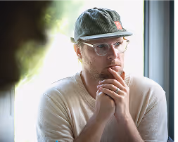 Thoughtful man wearing glasses and a cap, resting chin on clasped hands by a window.