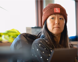 Woman with long dark hair wearing a rust-colored knit hat and gray polka dot sweater sitting indoors.