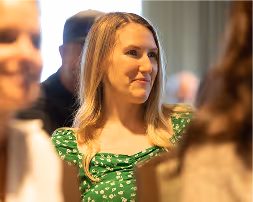 Young woman with long blonde hair in a green dress with white floral pattern, smiling indoors with blurred people around her.