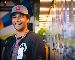 Smiling young man wearing a gray cap with a pink logo and a black shirt, standing next to a row of lockers indoors.