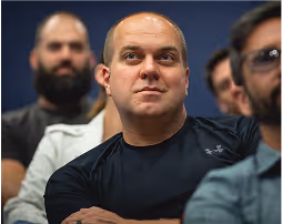 A bald man in a black shirt sitting among other people in an indoor setting.