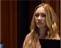 Young woman with long blonde hair and serious expression sitting indoors with dark curtains in the background.
