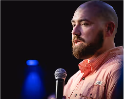 Bearded man in pink shirt speaking into a microphone with dark background and blue spotlight.