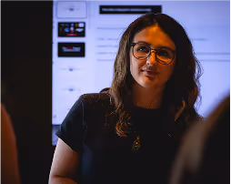 Woman with glasses standing in front of a screen with presentations in a dimly lit room.