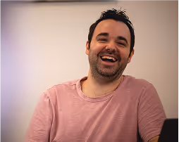 Man with dark hair and beard smiling and laughing, wearing a pinkish shirt against a plain background.