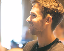 Profile of a smiling young man with short hair in a softly lit indoor setting.