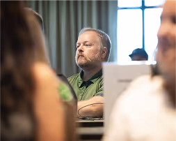 Man with a beard and green shirt attentively listening in a meeting room with blurred people in foreground.