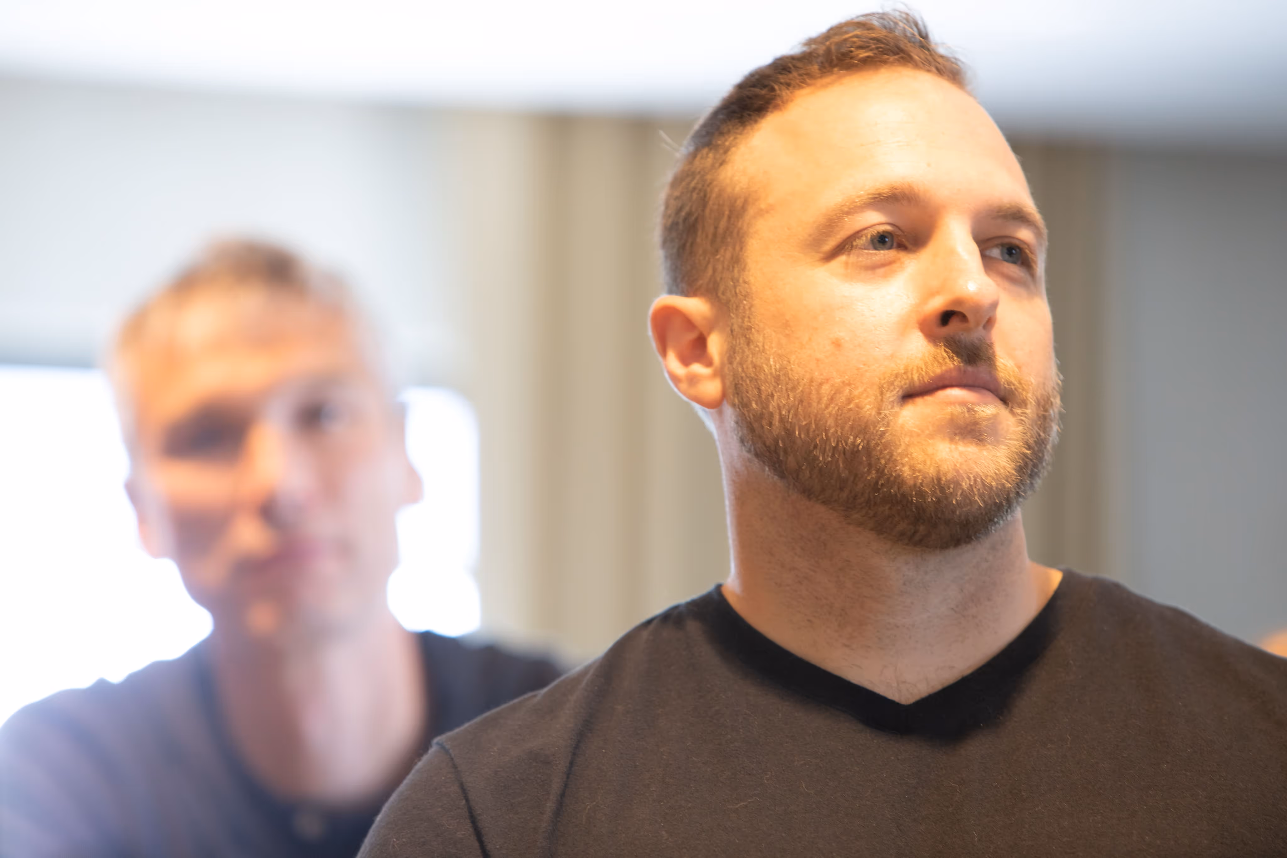 Bearded man in a black shirt looking to the side with a blurred man in the background indoors.