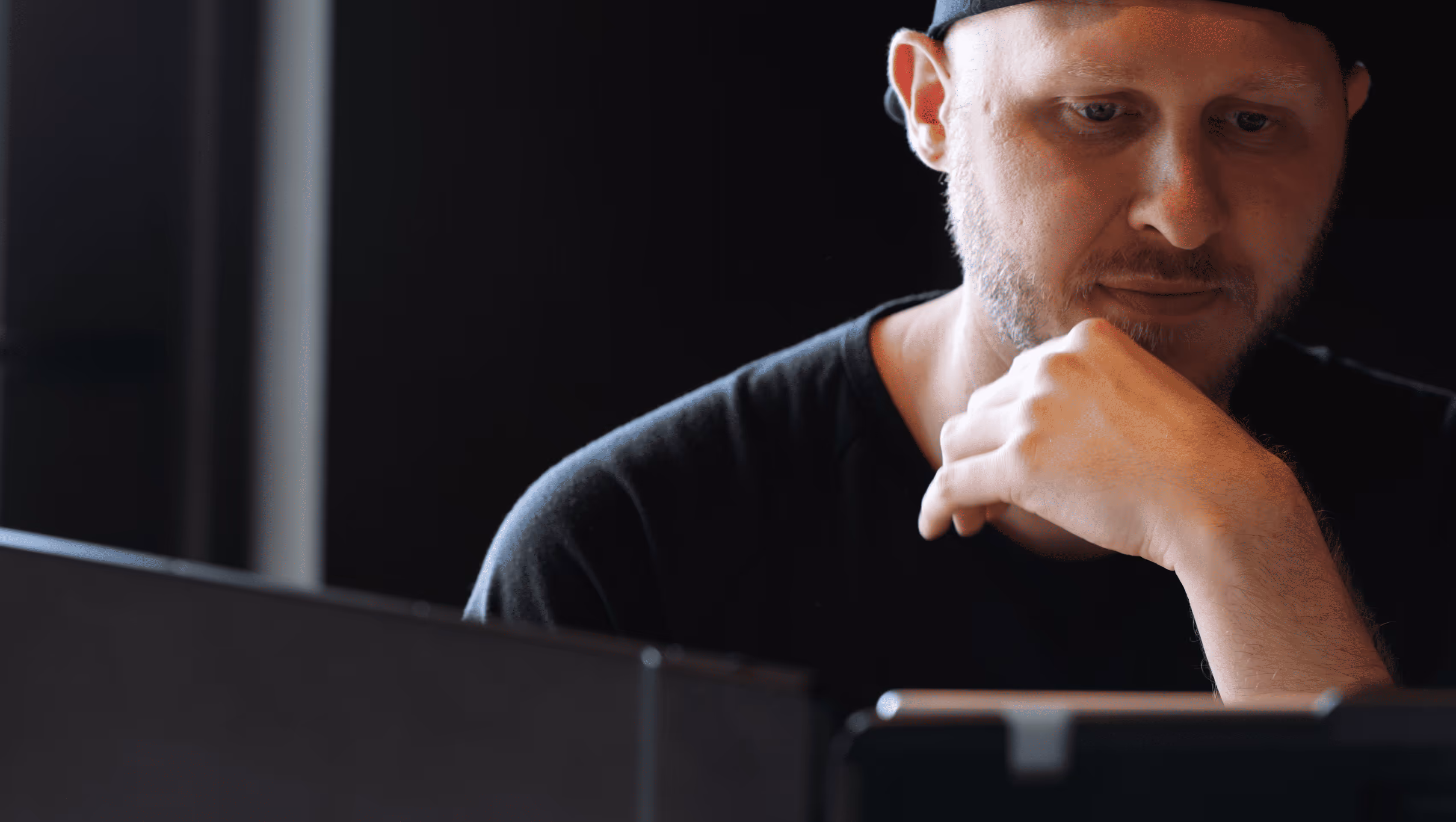 Man with a beard wearing a black shirt and cap looking thoughtfully at a computer screen.