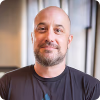 Bald man with a beard and mustache smiling, wearing a black shirt, in a softly lit indoor setting.